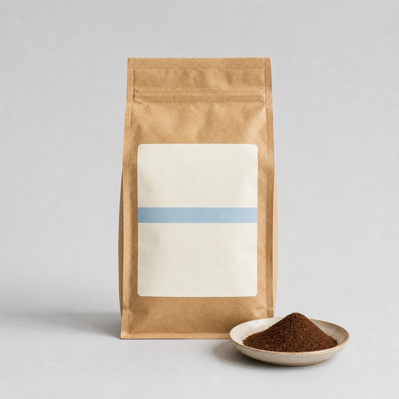 Brown kraft coffee bag with a pale blue stripe on the label, beside ground coffee on a saucer